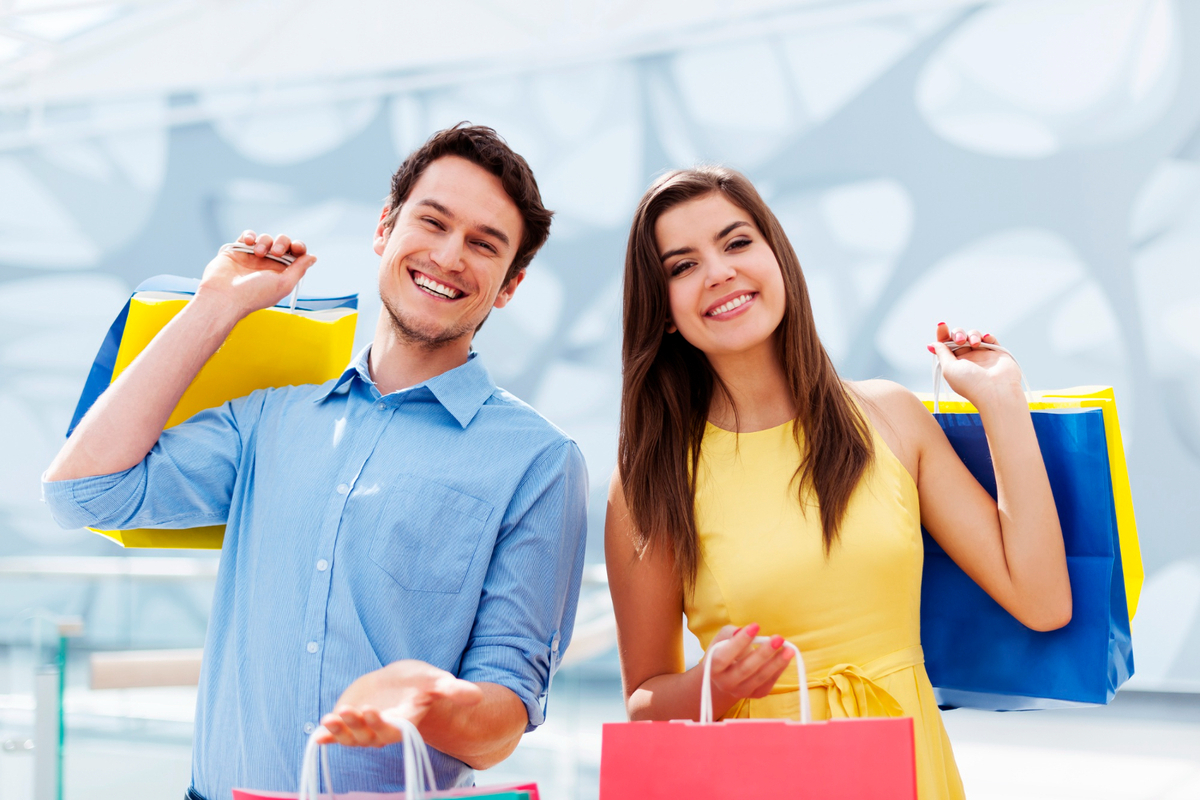 Homem e mulher lado a lado sorriem enquanto seguram sacolas de papel com boas compras e ao fundo uma parede decorada em azul claro